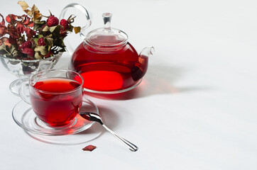 Breakfast with organic red hawthorn tea in transparent teapot, cup,  hawthorn berries, rose hip in sunlight with shadow on white wood board, copy space.