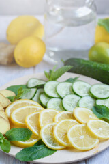 Obraz na płótnie Canvas Sassi water, cooking ingredients for making sassi drink: lemon, cucumbers, ginger root, mint on a round wooden tray. Light background. Close-up