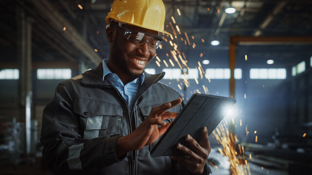 Professional Heavy Industry Engineer/Worker Wearing Safety Uniform and Hard Hat Uses Tablet Computer. Smiling African American Industrial Specialist Standing in a Metal Construction Manufacture.