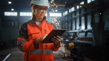 Professional Heavy Industry Engineer/Worker Wearing Safety Uniform and Hard Hat Uses Tablet Computer. Serious Successful Female Industrial Specialist Standing in a Metal Construction Manufacture.