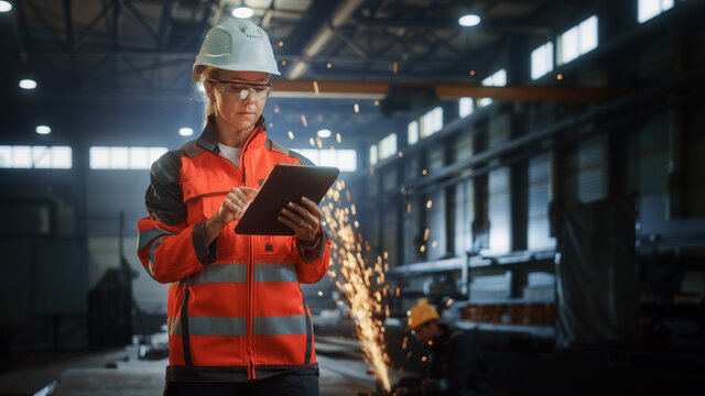 Professional Heavy Industry Engineer/Worker Wearing Safety Uniform and Hard Hat Uses Tablet Computer. Serious Successful Female Industrial Specialist Standing in a Metal Construction Manufacture.