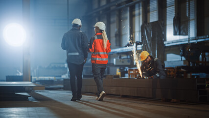 Fototapeta premium Two Heavy Industry Engineers Walk in Steel Factory, Use Tablet and Discuss Work. Industrial Worker Uses Angle Grinder in the Background. Black African American Specialist Talks to Female Technician.