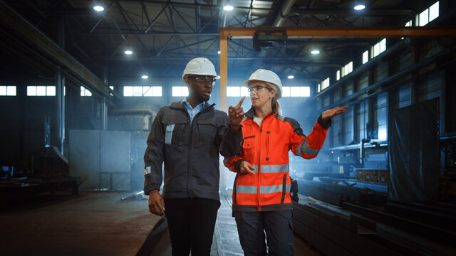 Two Heavy Industry Engineers in Hard Hats Walk in Steel Metal Manufacturing Factory and Have a Discussion. Black African American Industrial Specialist Talk to Female Technician in Manufacture.