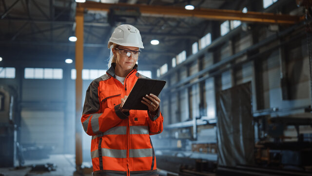 Professional Heavy Industry Engineer Worker Wearing Safety Uniform and Hard Hat, Using Tablet Computer. Serious Successful Female Industrial Specialist Walking in a Metal Manufacture Warehouse.