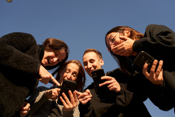 Obraz premium Low-angle of s group of friends using a smartphone and and laughing. He teaches them something fun on his phone. New normal lifestyle in the covid-19 season.