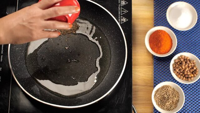 Female's hand dropping cumin seeds/whole Jeera in a heated pan with cooking oil. Closeup shot of black Chana  cumin  red chili powder  and salt in bowls with a non-stick pan for cooking - food maki...