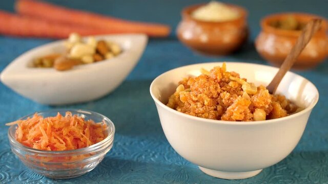 Dry fruits dropping in a bowl with grated carrots and carrot pudding on a table. Closeup shot of winter special Gajar Ka Halwa in a white ceramic bowl - traditional Indian sweet dish
