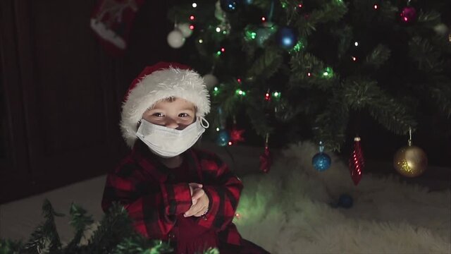 coronavirus, covid-19 in the new year. a little girl in a red Santa Hat and mask is sitting under the Christmas tree at home.