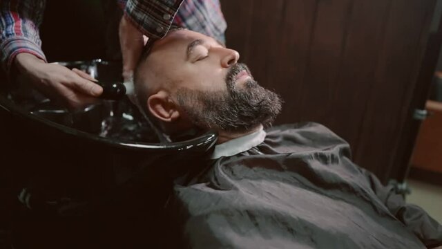Hairdresser washes the head of a bearded man in the sink.