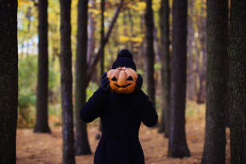 Young woman holding carved halloween pumpkin   in front of her face in forest. Funny Halloween concept 