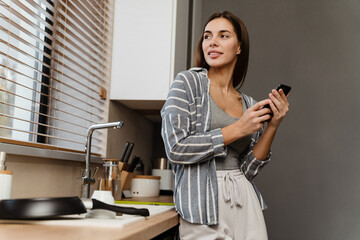 Charming happy woman smiling while using mobile phone in cozy kitchen