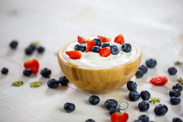 Bowl filled with yoghurt and berries on white table. Woody dish full of cream with strawberries and blueberries served as breakfast. Little sweet healthy fruit scattered around the brown stray.