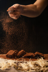 Close up of a hand is sprinkling cocoa powder over chocolate brownies on baking paper against black background