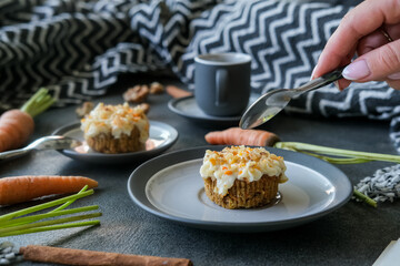 Close up of a hand with a spoon and delicious homemade carrot cupcake or muffin with ingredients and cup of coffee around on minimalist stone grey background