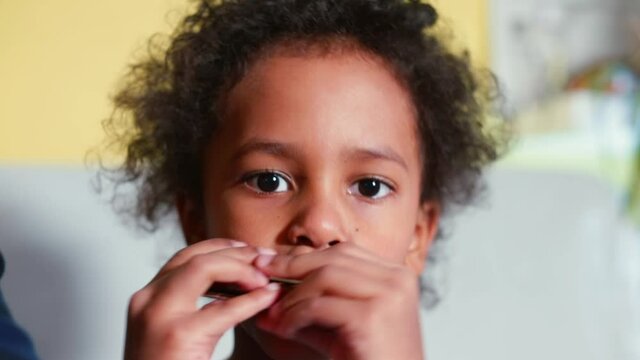 Portrait of a black child playing the harmonica. Hands move the device near the lips, makes sounds. A musical hobby in self-isolation.