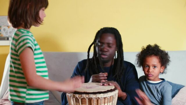Shows the technique of striking jambe with his hands. Training of technique and musical ear. Transfer of knowledge to the generation. Black father teaches children to play son's drum. 