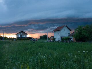 Fototapeta premium Stormy severe weather with arcus shaft and cumulonimbus clouds, cloudburst or shower and sunshine behind rain above settlement during summer evening.