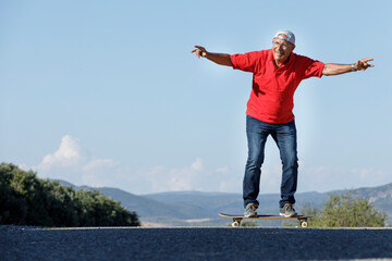Uomo settantenne con polo rossa, va nel suo skateboard stando in equilibrio in una strada di campagna - obrazy, fototapety, plakaty