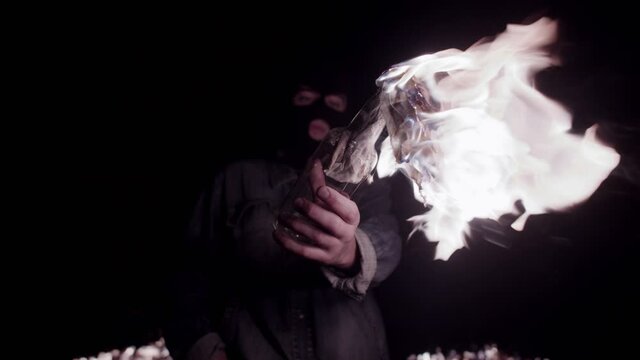 Man in denim jacket and balaclava shows camera molotov cocktail against background of burning earth. Confident guy directs glass bottle of gasoline to camera with rag burning in it