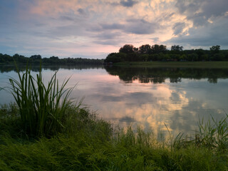 Landscape of Sava river in Poloj near Bosanski Brod, Bosnia and Herzegovina with green grassy and forested shore, calm water with reflection and beautiful clouds in sky during evening in summer