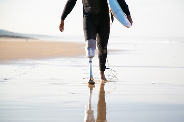 Unrecognizable surfer with leg prosthesis walking near sea. Strong man wearing black swimsuit, carrying surfboard and going along coastline. Artificial limb, lifestyle and extreme sport concept