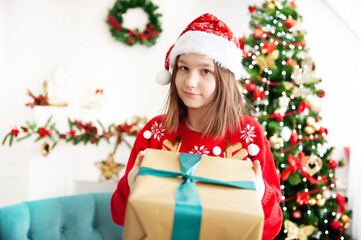 A young teenage girl in a Christmas cap and a red sweater with a large gift is sitting in a cozy chair next to the Christmas tree. Congratulation