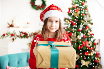 A young teenage girl in a Christmas cap and a red sweater with a large gift is sitting in a cozy chair next to the Christmas tree. Congratulation