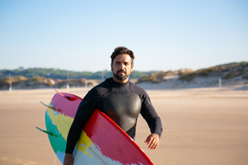 Attractive brunette surfer walking and sand beach with board. Caucasian bearded man holding surfboard and wearing wetsuit. Blurred background. Lifestyle, vacation and summer activity concept