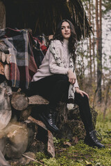 A woman of model appearance with long dark curly hair dressed in a white sweater scarf sits on a bench and poses against the background of neatly folded firewood