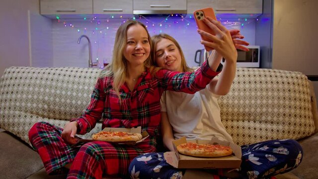 Beautiful cheerful girls in pajamas speaking with their friends by video call. Stylish young women sitting on the sofa with piazza, laughing and taking a photos. Social distance concept. Covid-19.