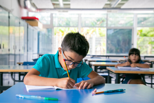Focused young boy in glasses sitting at desk and writing in copybook in class. Copy space. Education or back to school concept