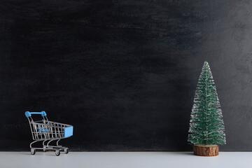 Shopping cart and miniature Christmas tree on black background. New year shopping. Copy space