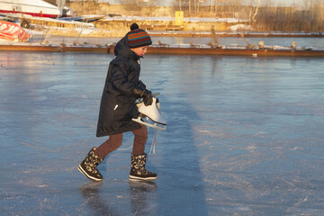 A boy walks on a frozen river in winter and carries white skates in his hands to ride on the ice.
