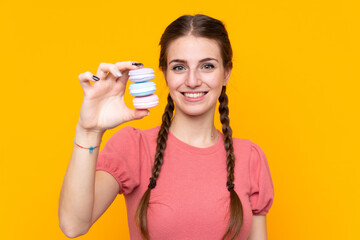 Young woman over isolated yellow background holding colorful French macarons and with happy expression