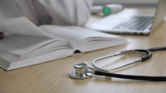 Work space of a doctor who reads books of medicine and research additional information in internet on a blurred background. Focus on stethoscope