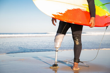 Unrecognizable male surfer standing with surfboard on sea beach. Cropped view of amputee with...