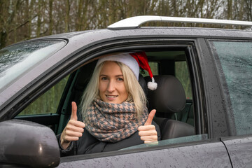 Young blonde woman in a Santa Claus hat is sitting at the wheel of a car and shows her hands a Thumbs up gesture