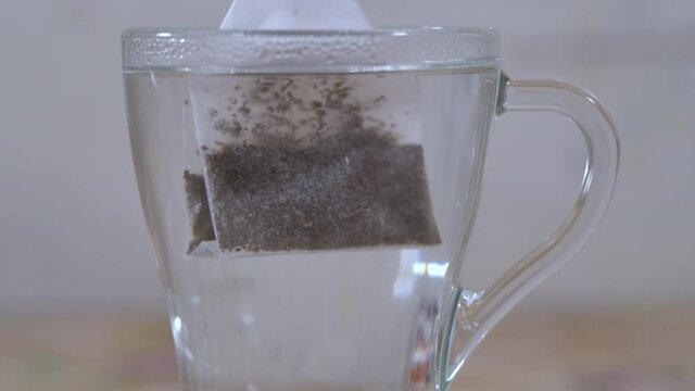 Slow Motion Close Up Glass Cup With Black Tea Bag. Making Morning Tea. Putting Tea Bag Into Glass Cup Full of Hot Water Slow Motion Closeup.