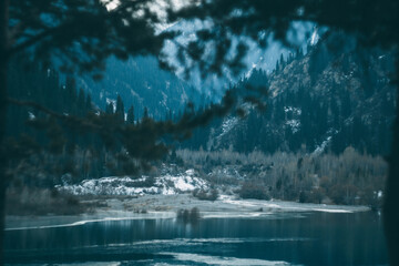 Blue mountain frozen lake in winter. Branches of trees in the foreground and mountains in the...