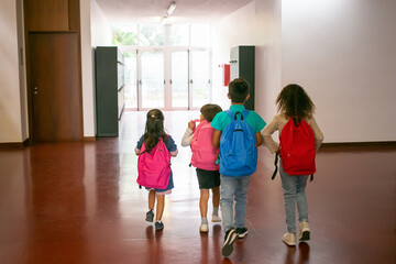 Group of children with colorful backpacks walking in school corridor to entrance. Back view, full length. Education or back to school concept