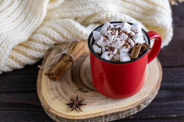 Hot chocolate with marshmallows, cinnamon, star anise - a winter hot spicy drink in a red cup on a wooden cut, with a white knitted scarf on a wooden background