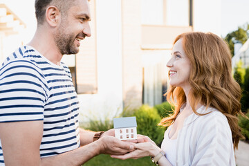 Side view of happy couple looking at each other while holding statuette of house on blurred background