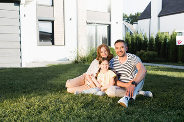 Happy family looking at camera while sitting on lawn near modern house
