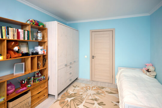 Cozy bright children's room with blue walls pastel carpet on gray wooden floor wooden bookshelf door bed and wooden wardrobe. Real estate apartment.