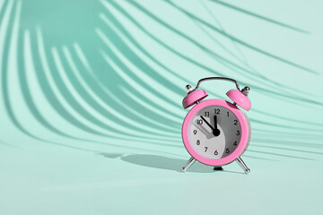 Pink alarm clock on light blue, turquoise pastel background with a shadow of palm leaf. Summertime,holiday, travel. Minimalism. Copy space.
