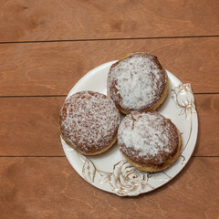 Three donuts, sprinkled with powdered sugar, lie on a plate. The dishes are white with a relief golden pattern in the form of rose flowers. The background is beige boards. Top view.
