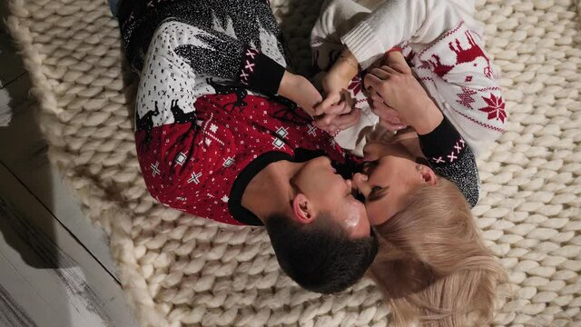 A couple in love in Christmas sweaters lies on the floor on a knitted blanket at home. The view from the top.