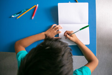 Black haired schoolboy sitting at desk, holding pen and writing in notebook. Top view. Education or back to school concept