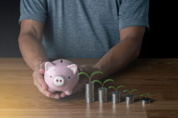The piggy bank is in the hand of the man and a pile of coins with a treetop on top, Money saving concept.