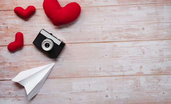 Wooden flat lay background for Valentines day holiday on February 14. White origami paper airplane, red stuffed plush toy hearts, and black with silver old retro film camera.